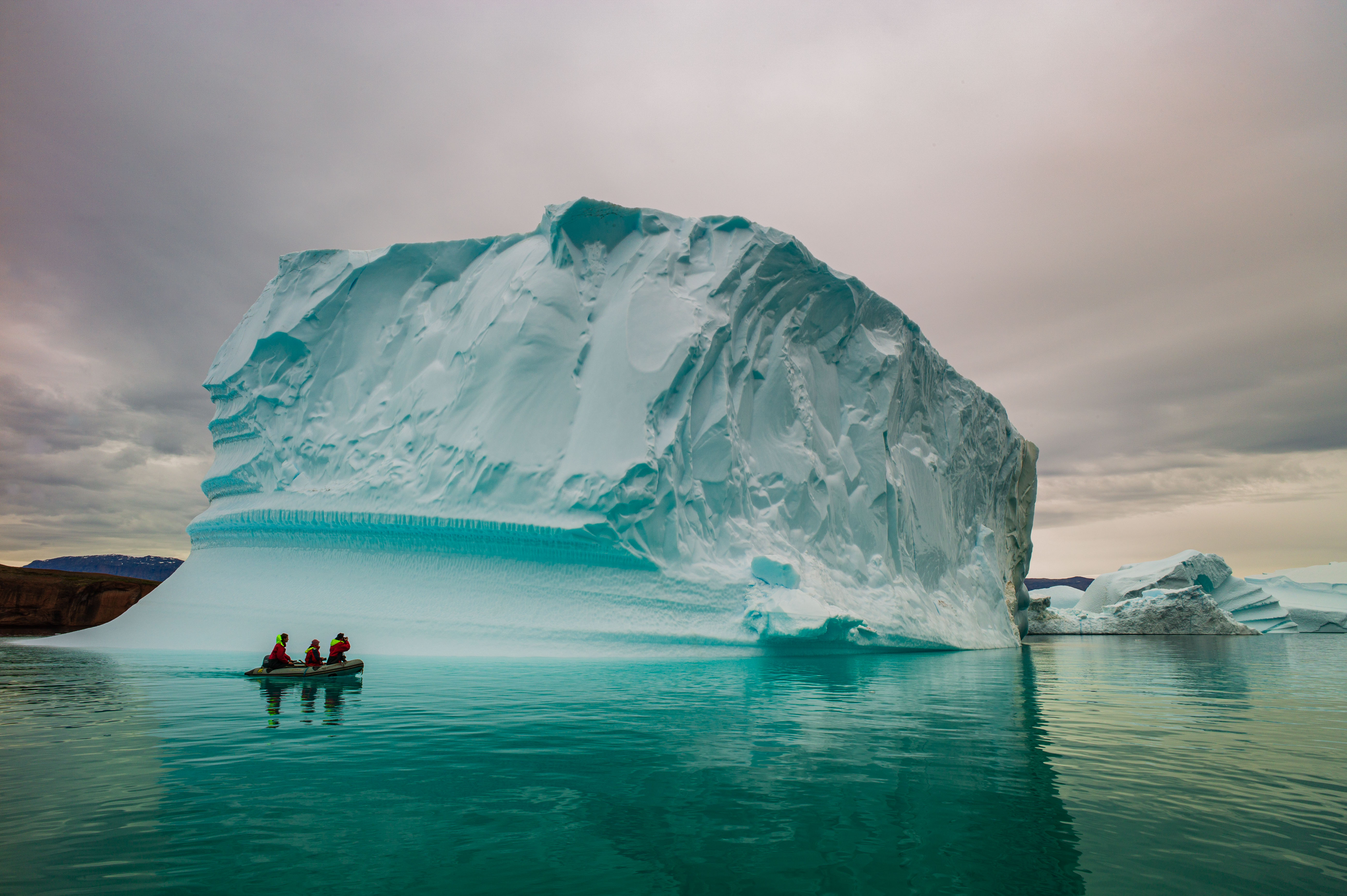 Three expedition members in a Zodiac beside a monumental iceberg in Scoresby Sund, East Greenland.