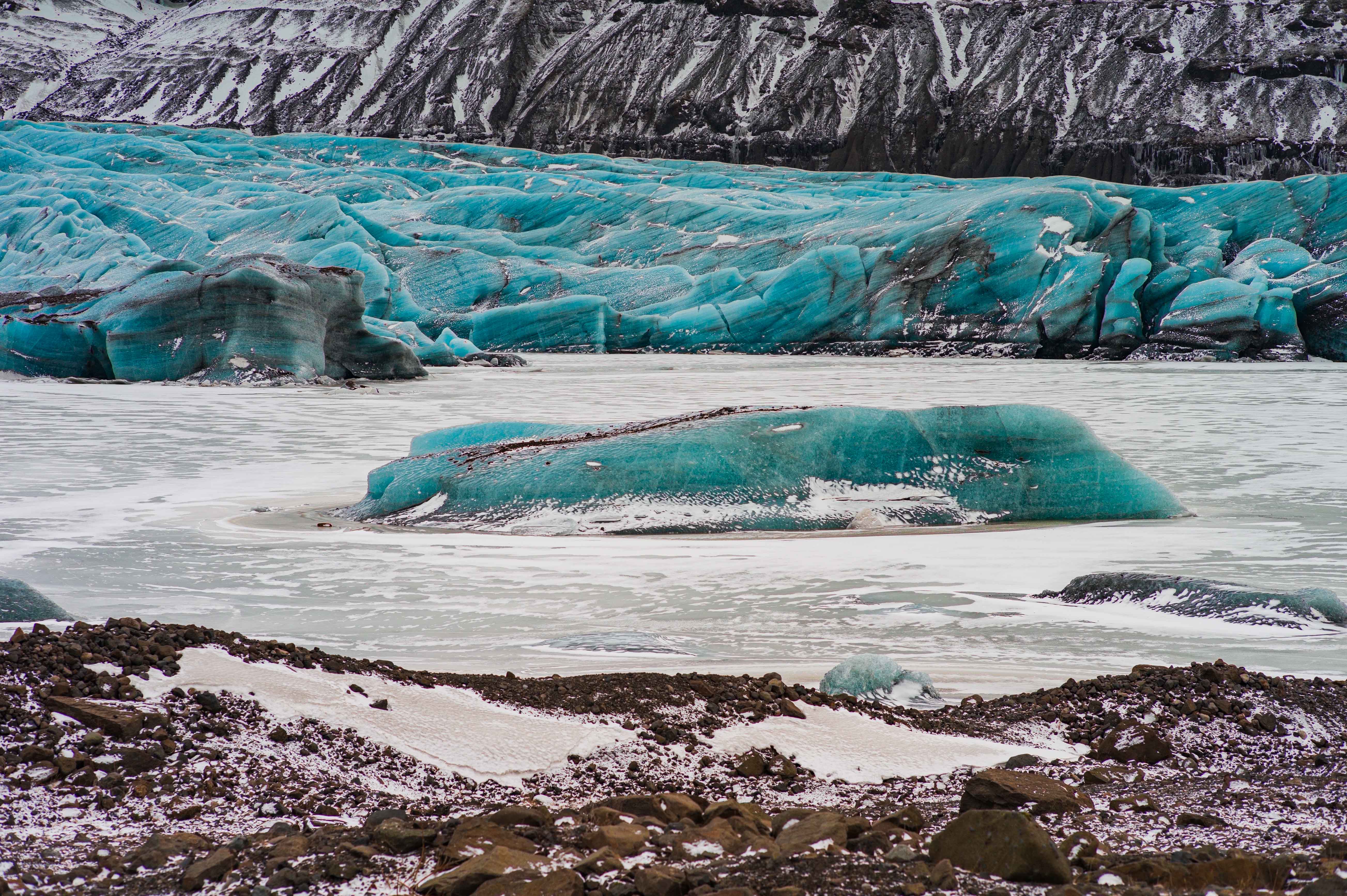 Blue glacier ice and detached ice blocks sit in pale meltwater at Fjallsárlón, framed by steep dark mountains and snow-covered slopes.
