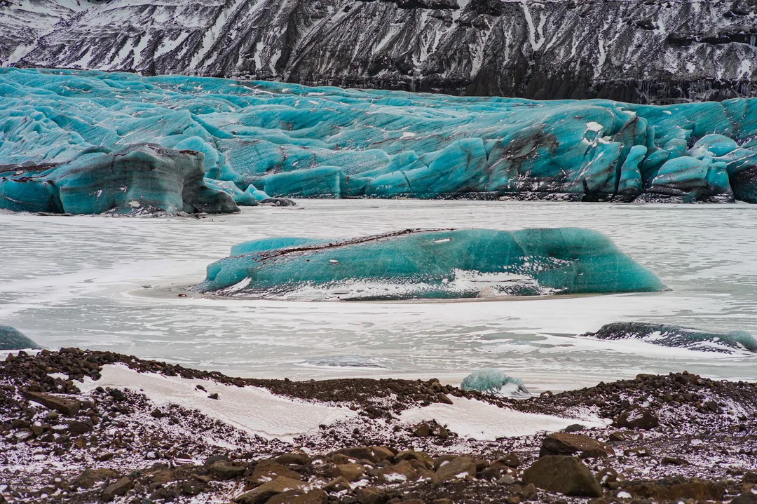 Blue glacier ice and detached ice blocks sit in pale meltwater at Fjallsárlón, framed by steep dark mountains and snow-covered slopes.