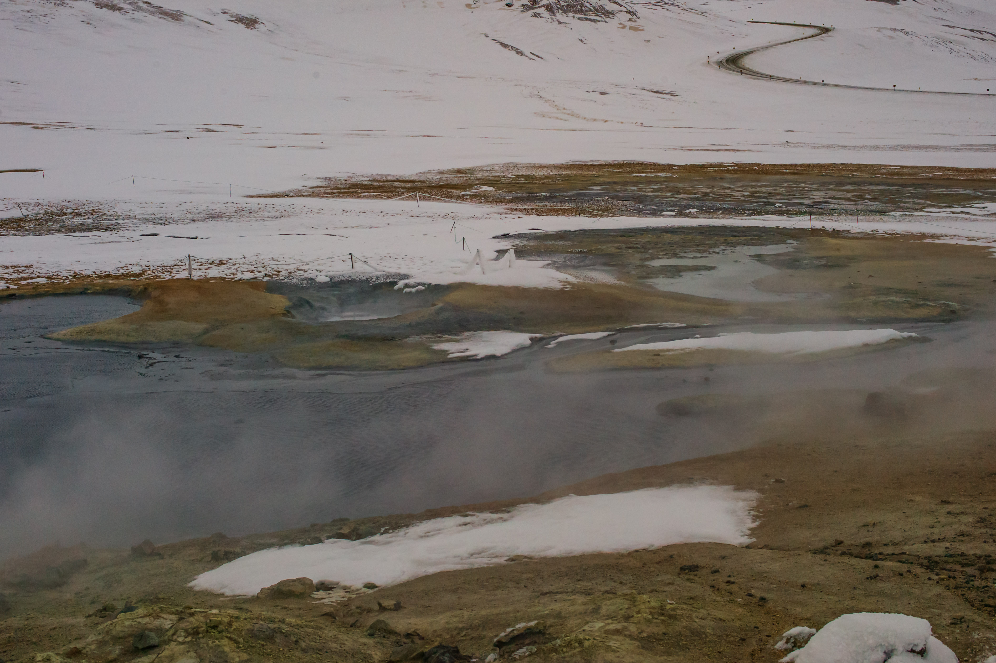 Wide winter view across the Hverir geothermal field, with shallow steaming pools, mineral-stained ground, patches of snow and a winding road on the pale mountainside beyond.