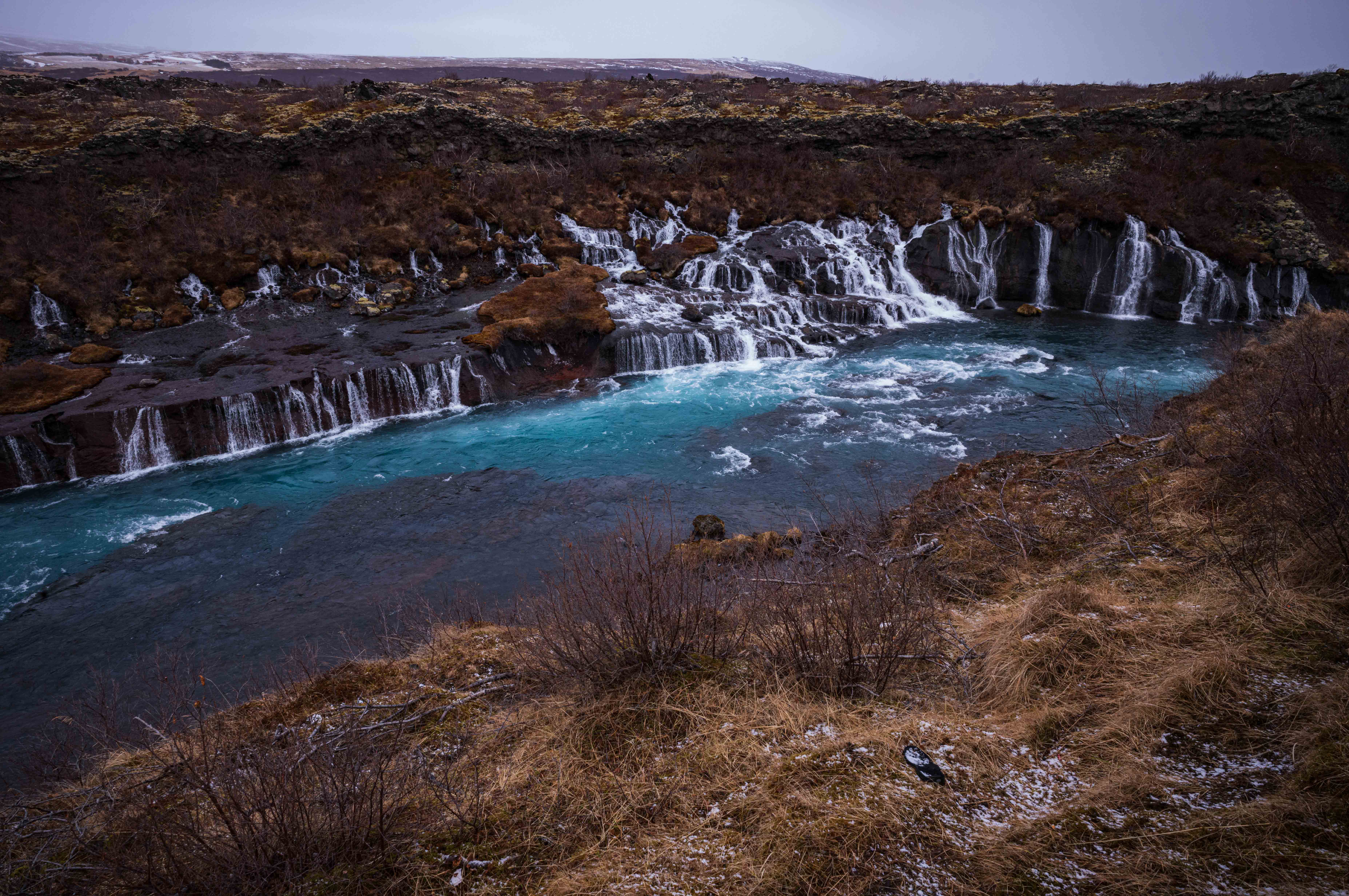 Turquoise river below a dark lava field at Hraunfossar, with numerous narrow waterfalls emerging through the rocky bank into the water.