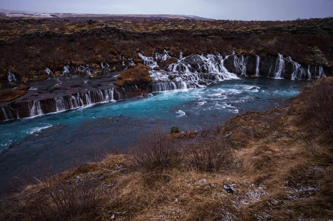 Turquoise river below a dark lava field at Hraunfossar, with numerous narrow waterfalls emerging through the rocky bank into the water.