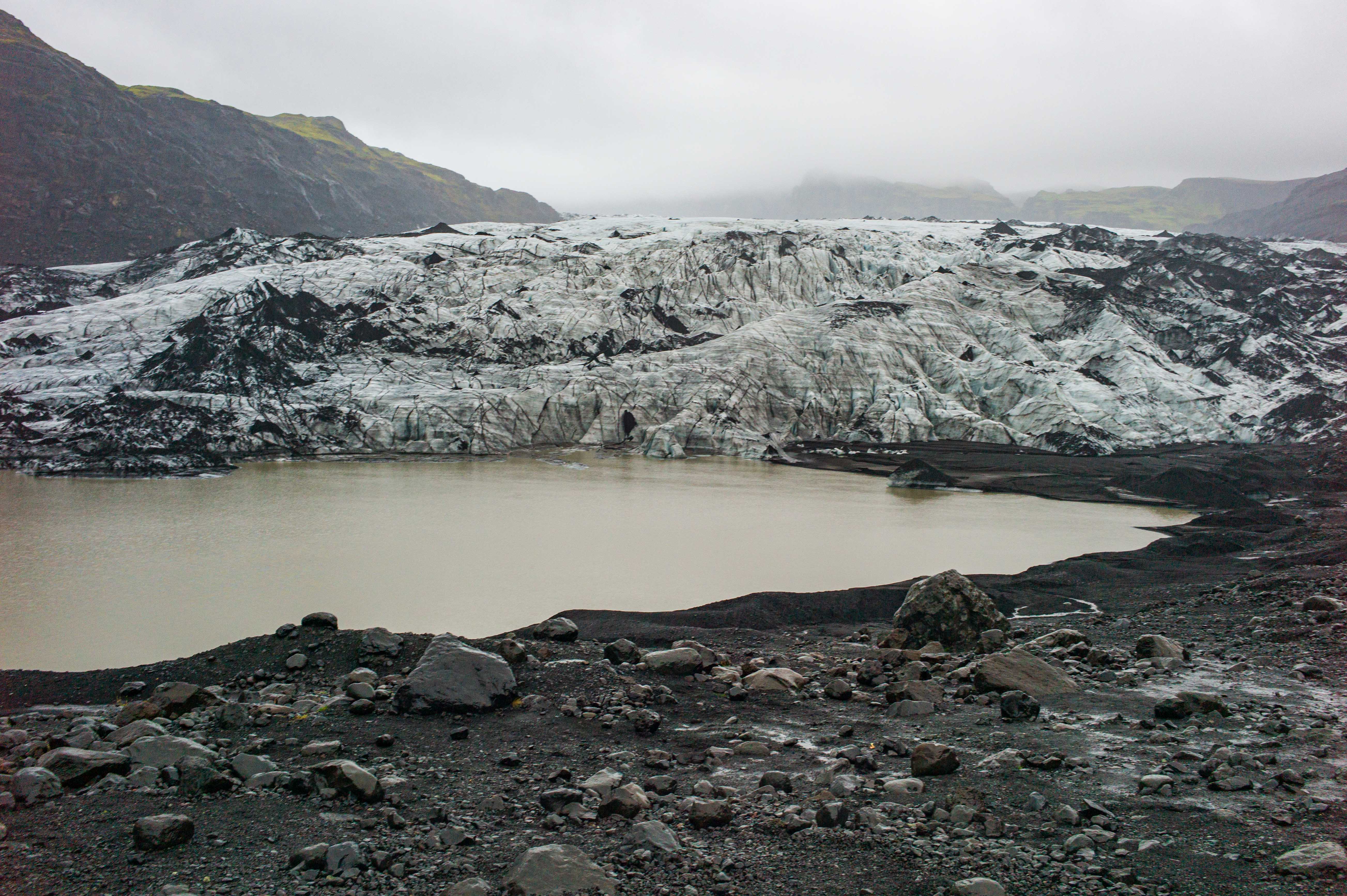 Wide view of the debris-streaked Sólheimajökull glacier behind a pale glacial lagoon, surrounded by dark volcanic sediment and rocky moraine.