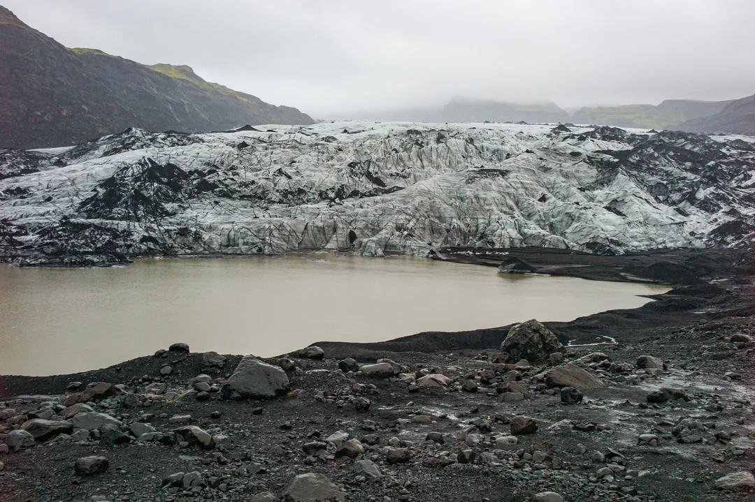 Wide view of the debris-streaked Sólheimajökull glacier behind a pale glacial lagoon, surrounded by dark volcanic sediment and rocky moraine.