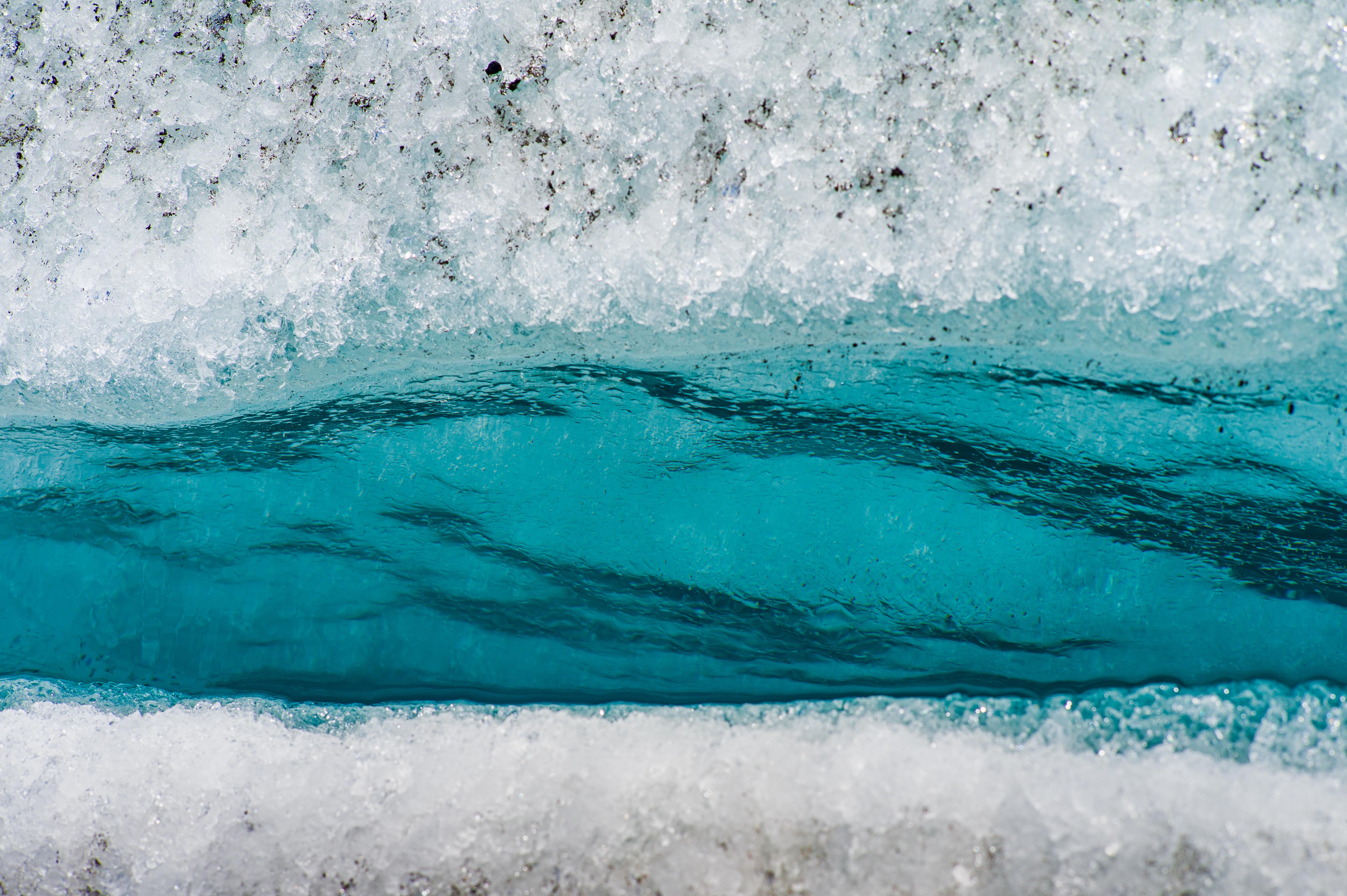 Close-up of vivid turquoise meltwater running through a narrow fracture in white glacier ice, with dark sediment embedded in the surrounding surface.