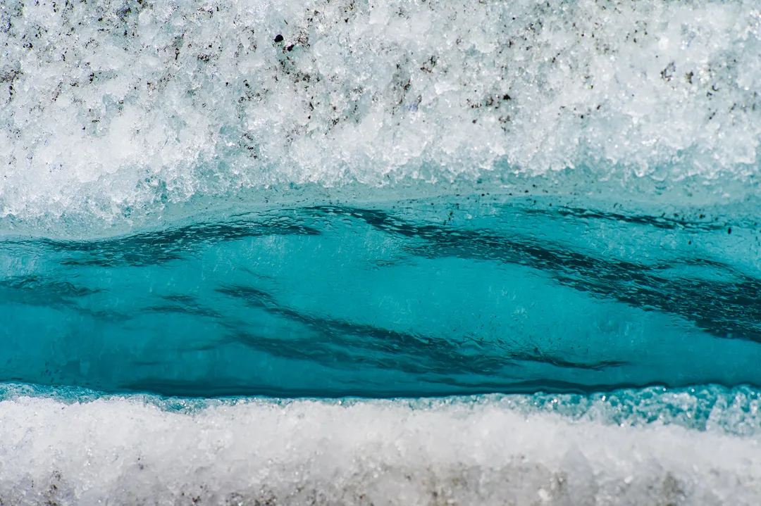 Close-up of vivid turquoise meltwater running through a narrow fracture in white glacier ice, with dark sediment embedded in the surrounding surface.