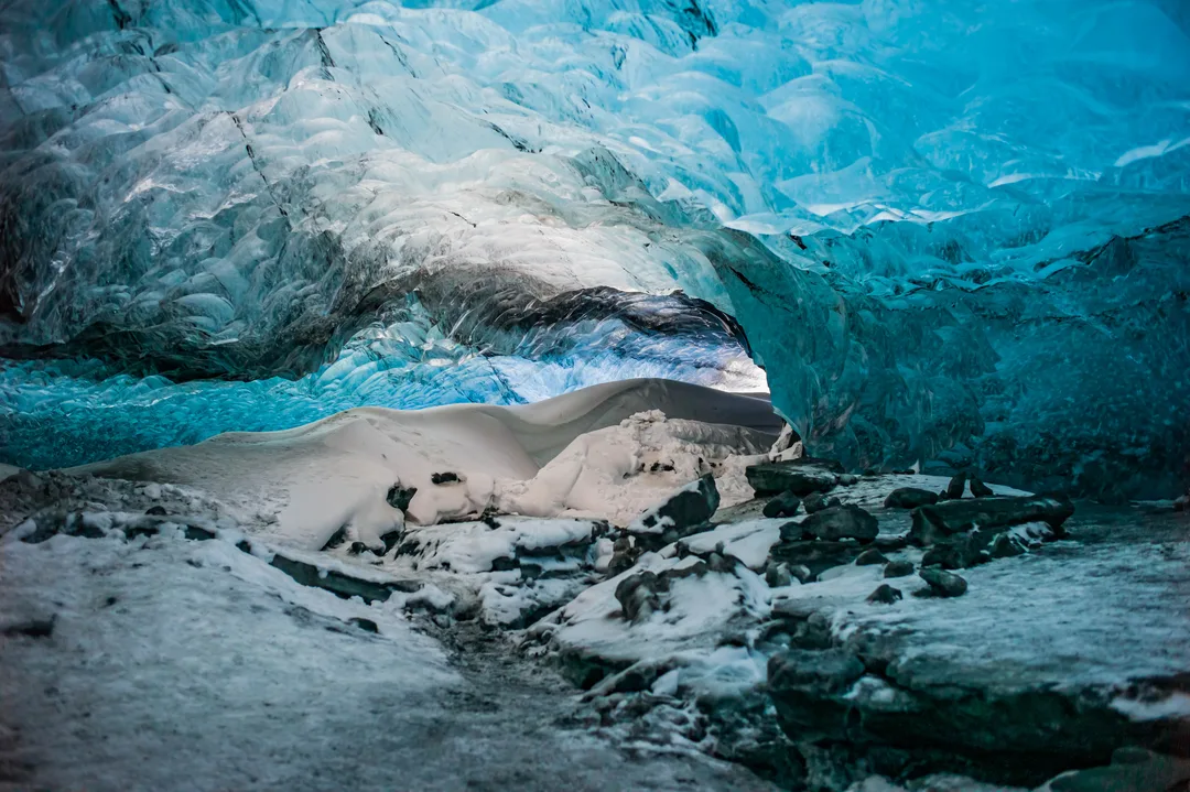 Blue translucent ice forms a low arched glacier cave above snow-covered ground and dark rocks, with layered ice textures visible across the ceiling and walls.
