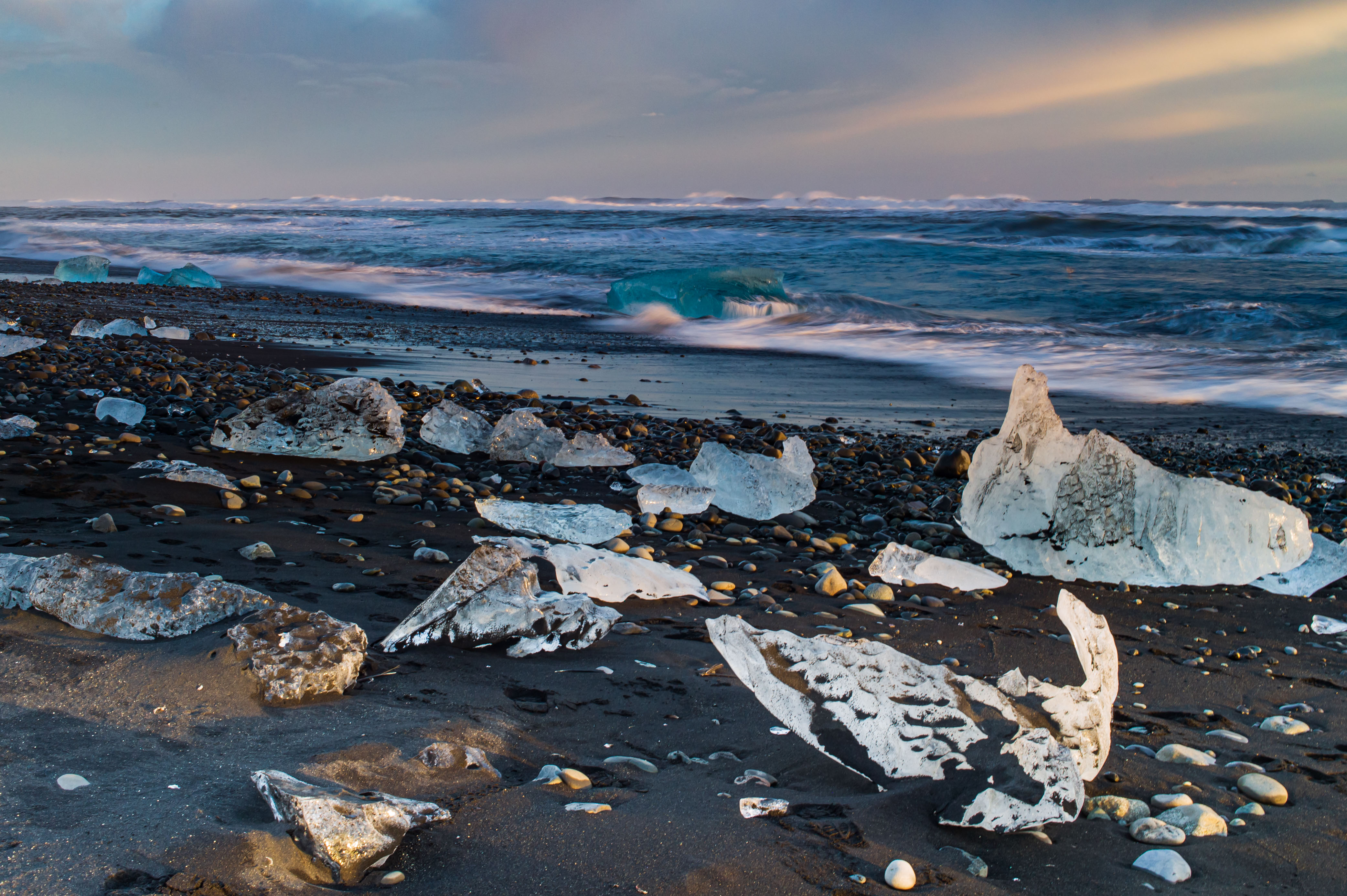Scattered clear and blue glacier-ice fragments resting on a black volcanic beach, with waves breaking behind them beneath a pale winter sky.