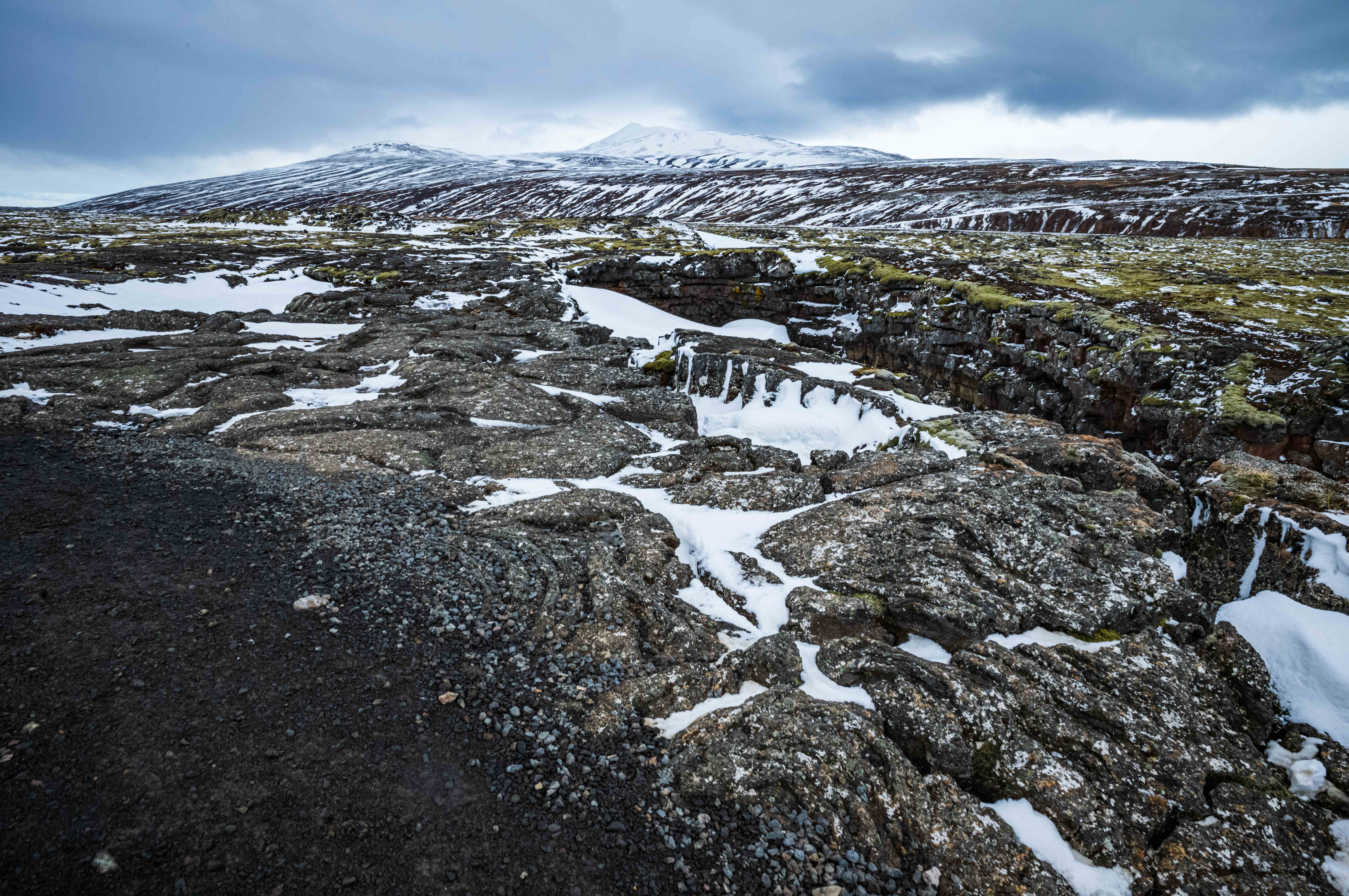 Wide view of fractured dark lava and snow-covered depressions near the entrance to Víðgelmir, with a broad volcanic plateau and snow-streaked mountains beyond.