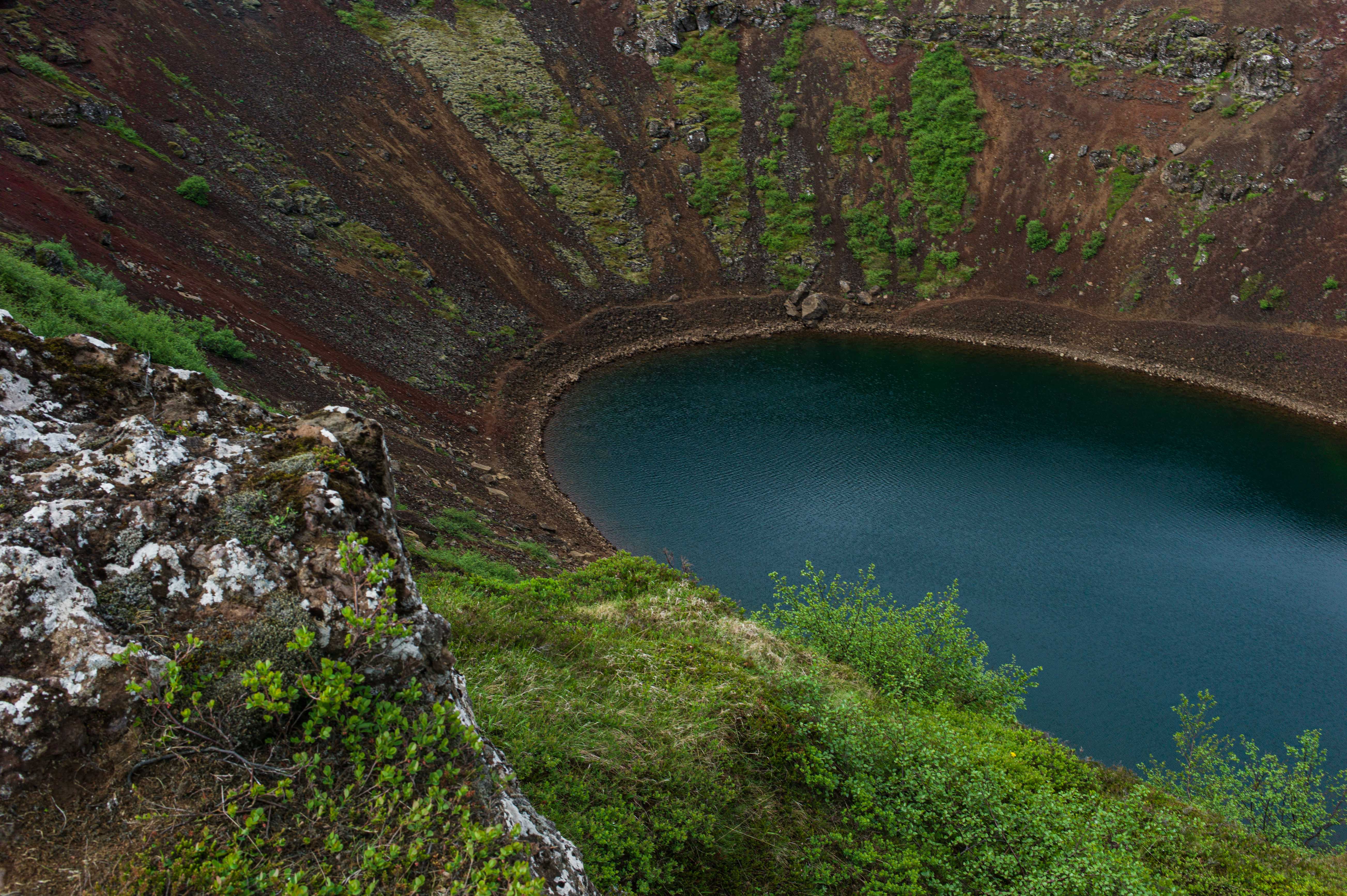 Deep blue-green crater lake surrounded by steep red-brown volcanic slopes streaked with green vegetation at Kerið in South Iceland.
