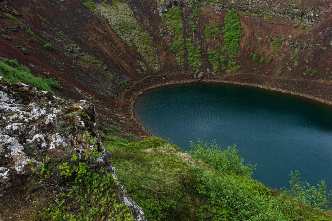 Deep blue-green crater lake surrounded by steep red-brown volcanic slopes streaked with green vegetation at Kerið in South Iceland.