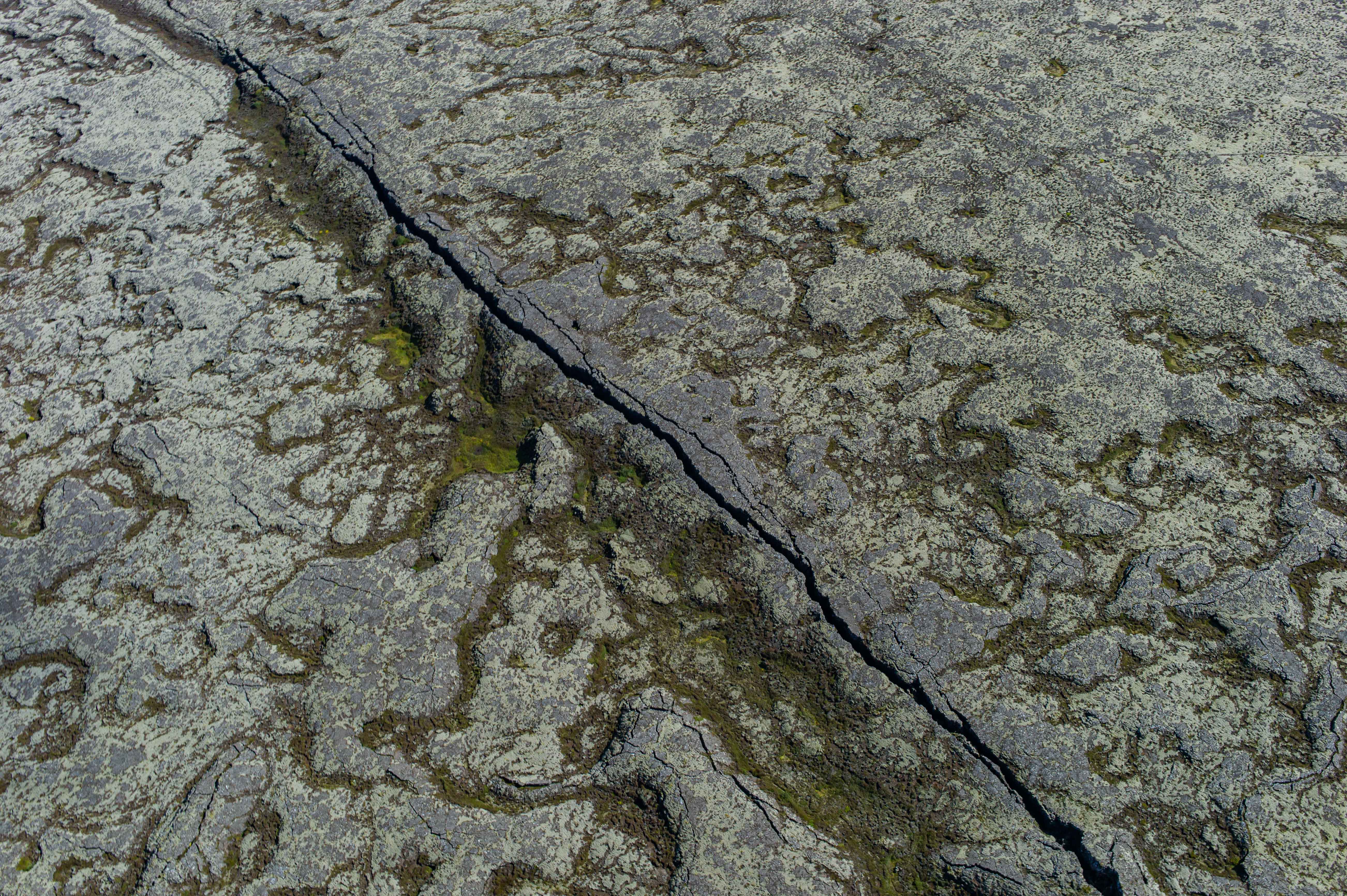 Aerial view of a narrow dark fissure running diagonally through pale, lichen-covered volcanic rock in Southwest Iceland.