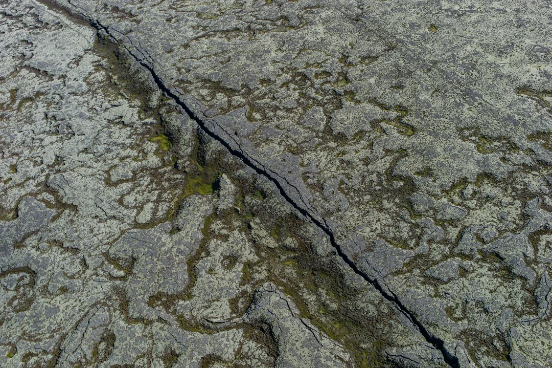Aerial view of a narrow dark fissure running diagonally through pale, lichen-covered volcanic rock in Southwest Iceland.