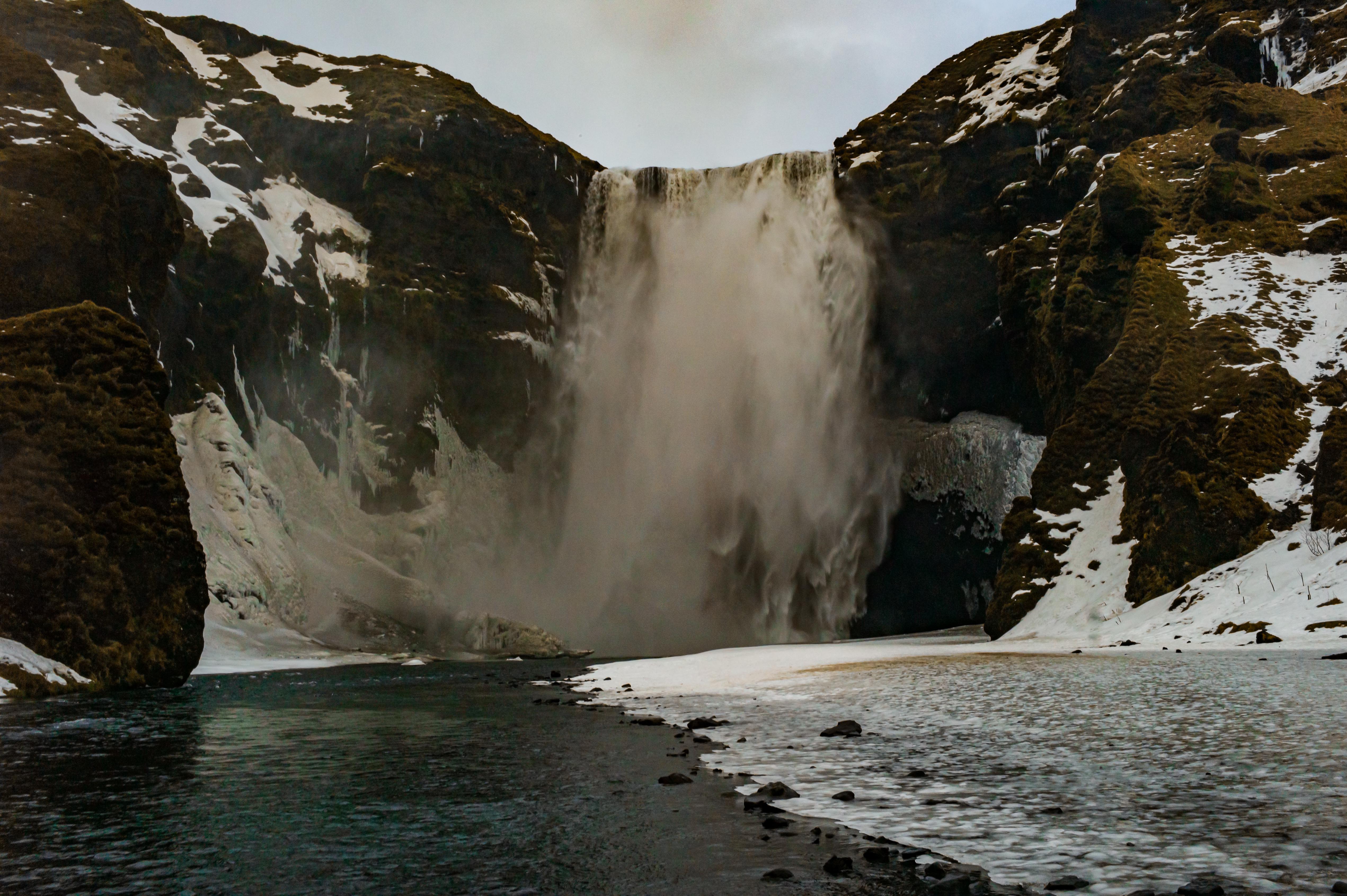 Skógafoss waterfall plunging between dark moss-covered cliffs, with snow and ice lining the river and banks below.