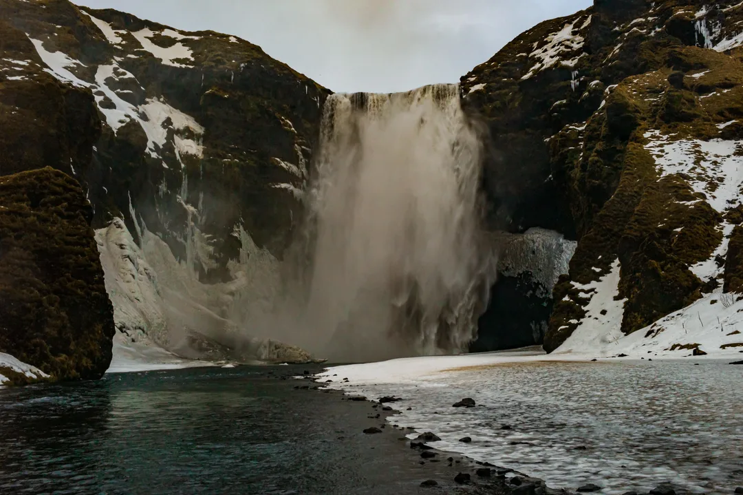 Skógafoss waterfall plunging between dark moss-covered cliffs, with snow and ice lining the river and banks below.