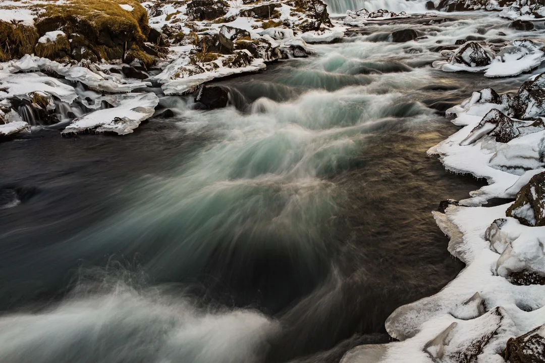 Fast blue-grey water rushing over low volcanic ledges at Fossálar, with snow-covered rocks and a long dark cliff rising behind the river.