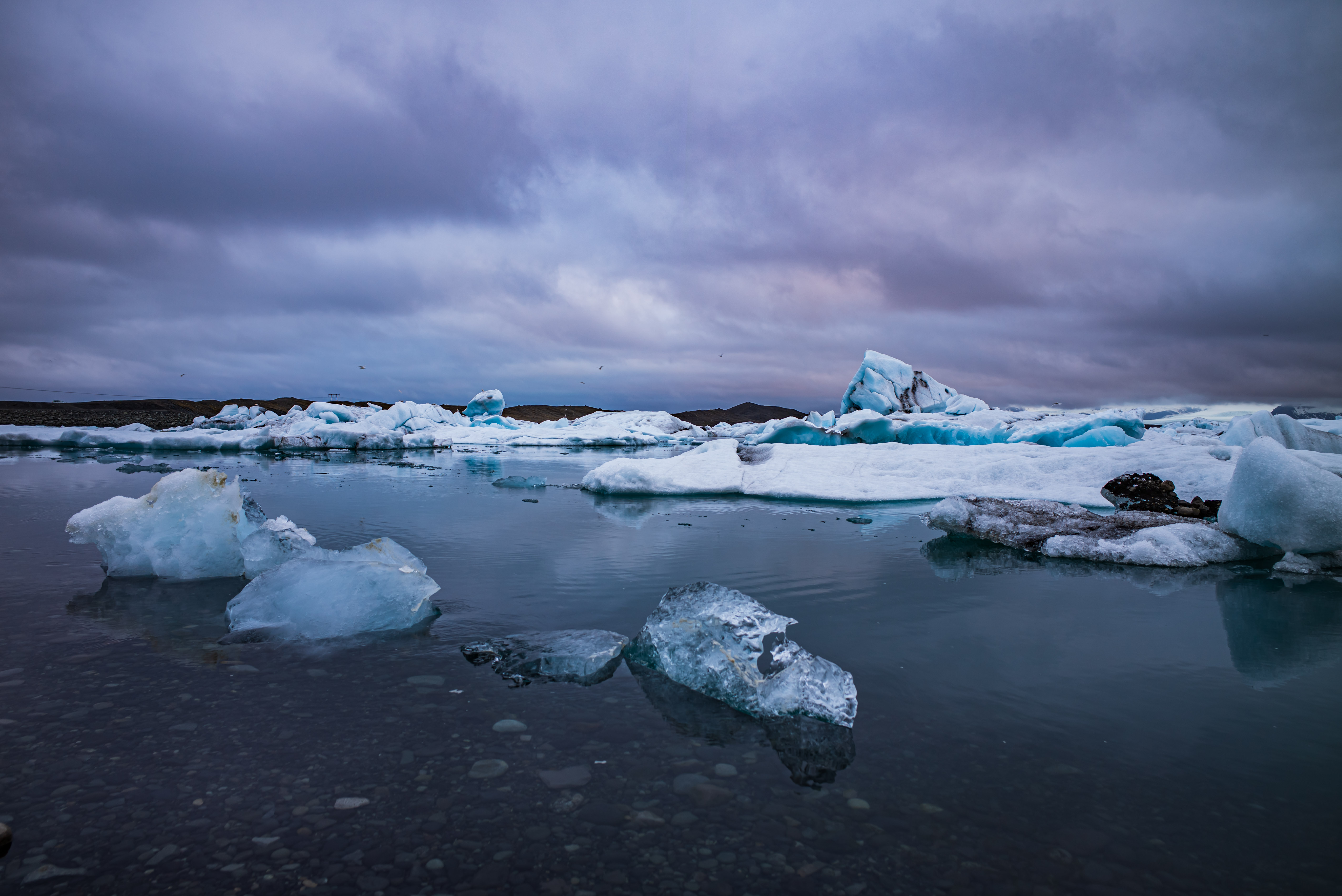 Blue-white icebergs floating in dark open water beneath a heavy grey sky at Jökulsárlón Glacier Lagoon, with smaller ice fragments in the foreground.