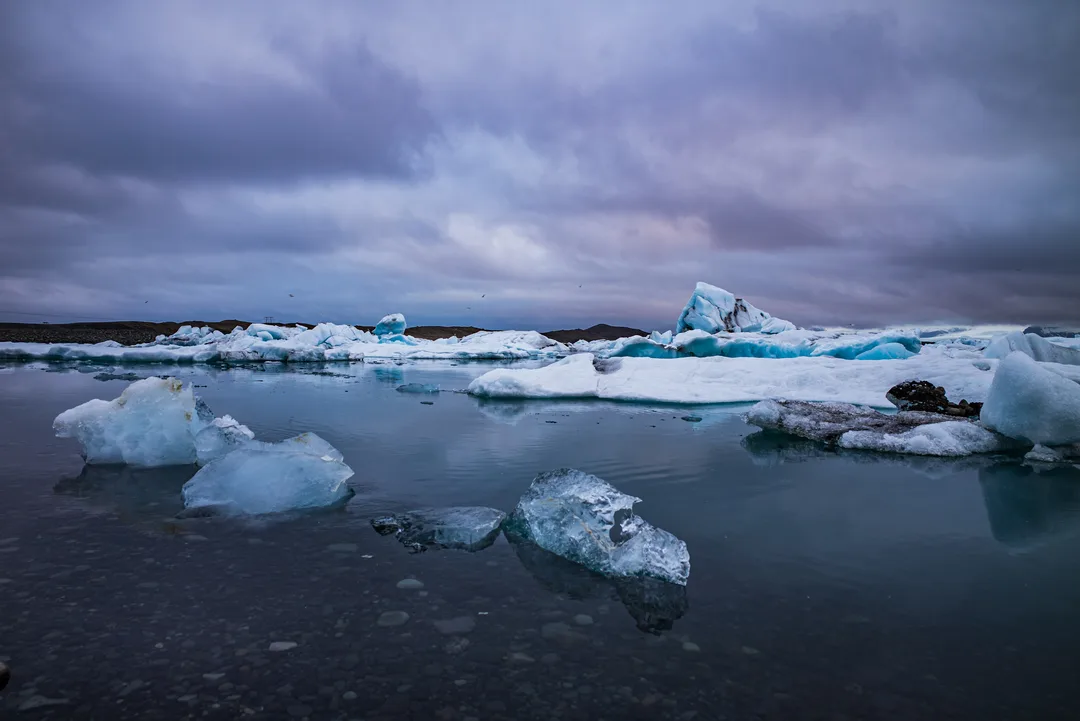 Blue-white icebergs floating in dark open water beneath a heavy grey sky at Jökulsárlón Glacier Lagoon, with smaller ice fragments in the foreground.
