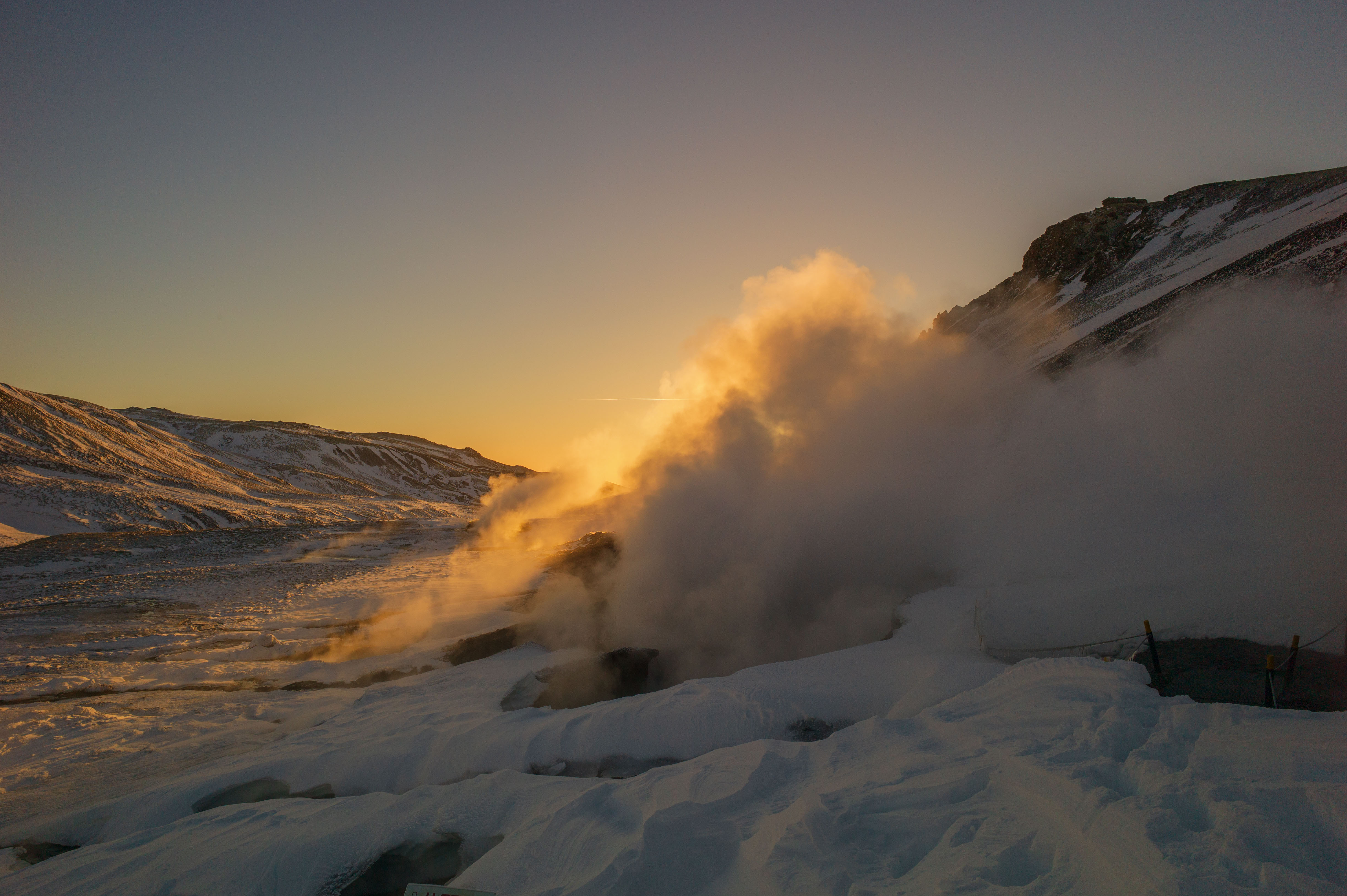Dense golden steam rising through a snow-covered geothermal valley at sunset near Reykjadalur, with dark volcanic slopes behind.